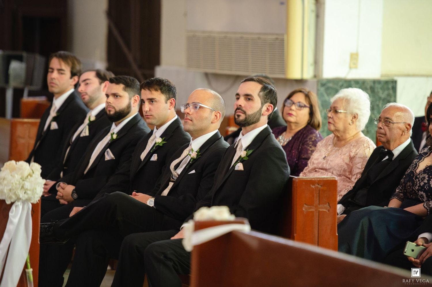 Groomsmen and family seated in church pews during a wedding ceremony, wearing black suits with white boutonnieres.