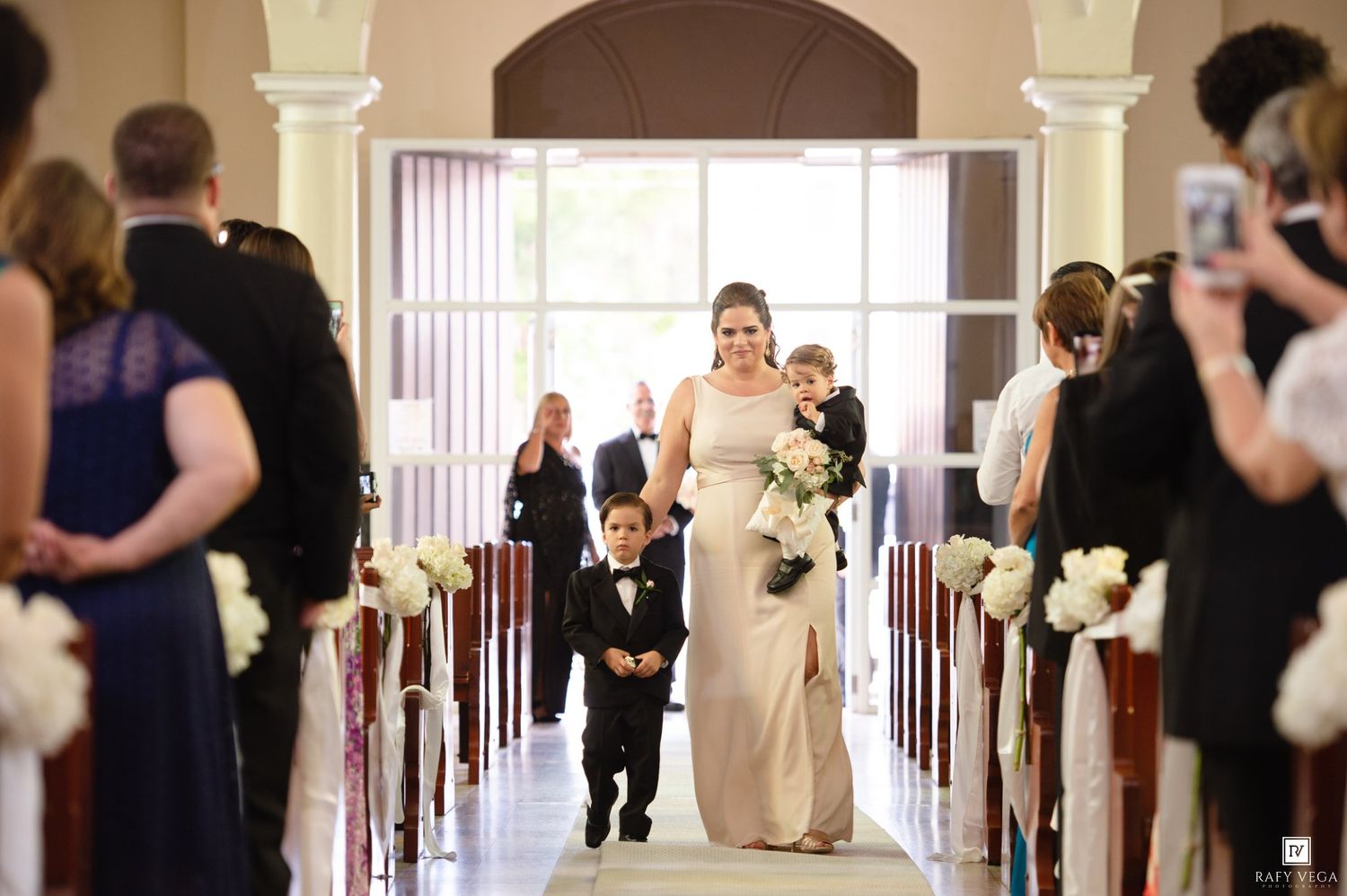 Bride walking down church aisle with young ring bearer, guests watching, white floral pew decorations.