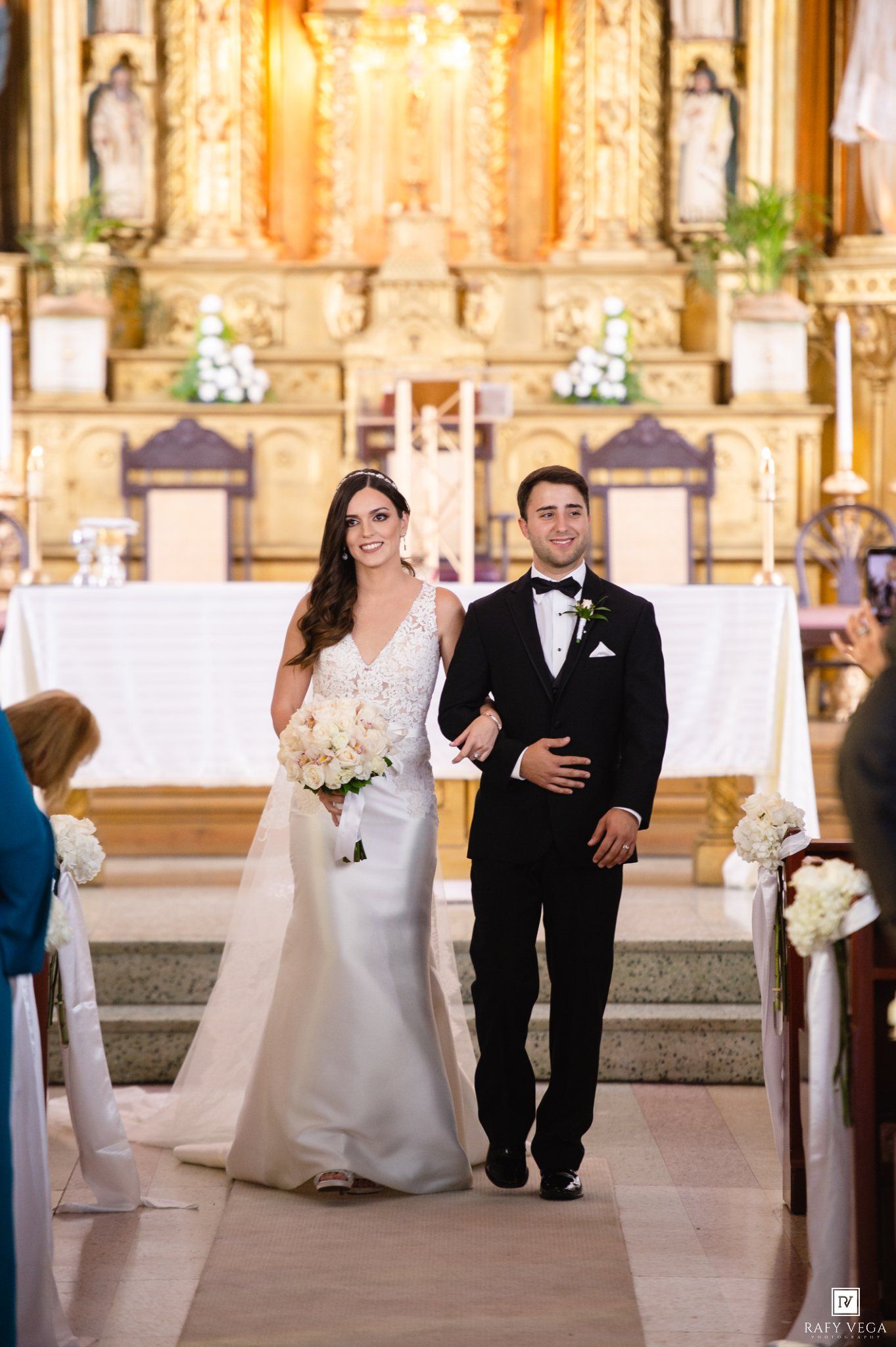 Bride and groom walk down church aisle after wedding ceremony, bride holding white bouquet in elegant gown.