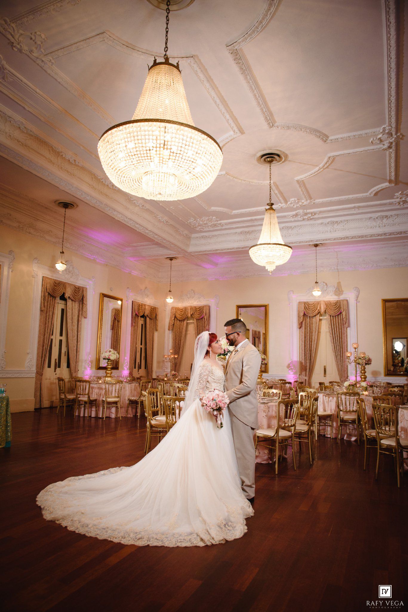 Bride and groom pose in elegant ballroom with crystal chandeliers, gold chairs, and soft pink uplighting on wedding day.