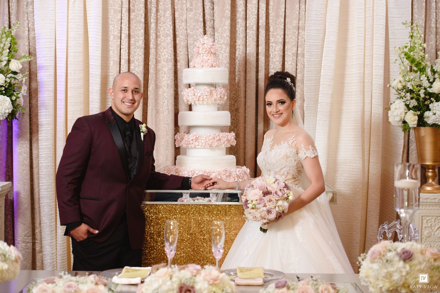 Bride and groom cutting a tall elegant white wedding cake at a luxury reception with floral decorations.