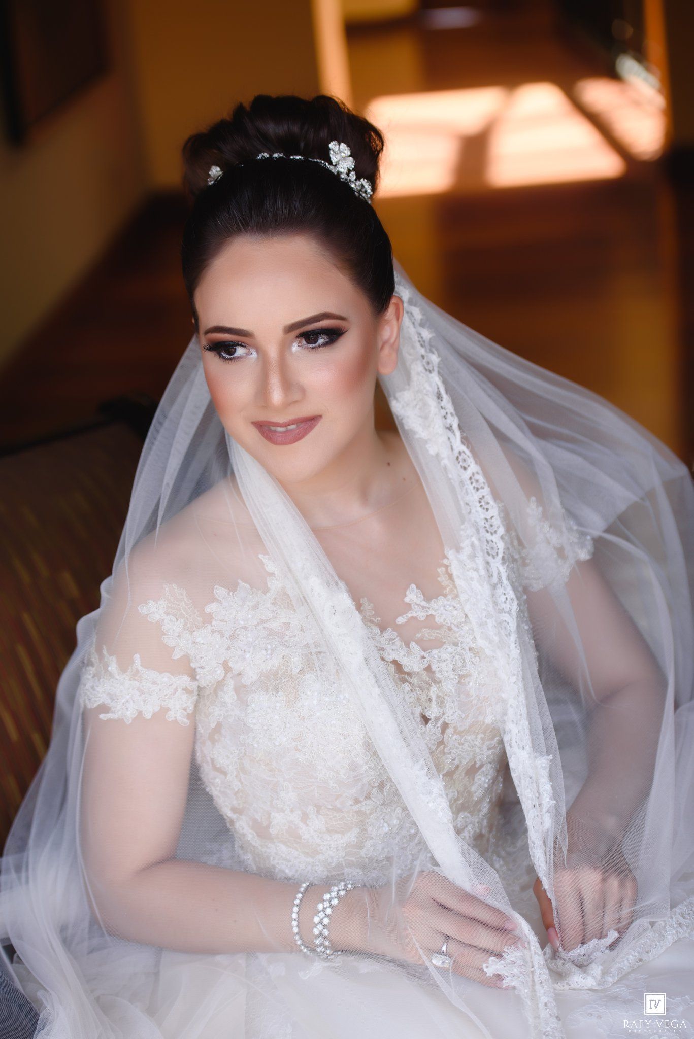 Elegant bride in lace wedding gown and veil wearing a tiara, with glamorous makeup and updo hairstyle.