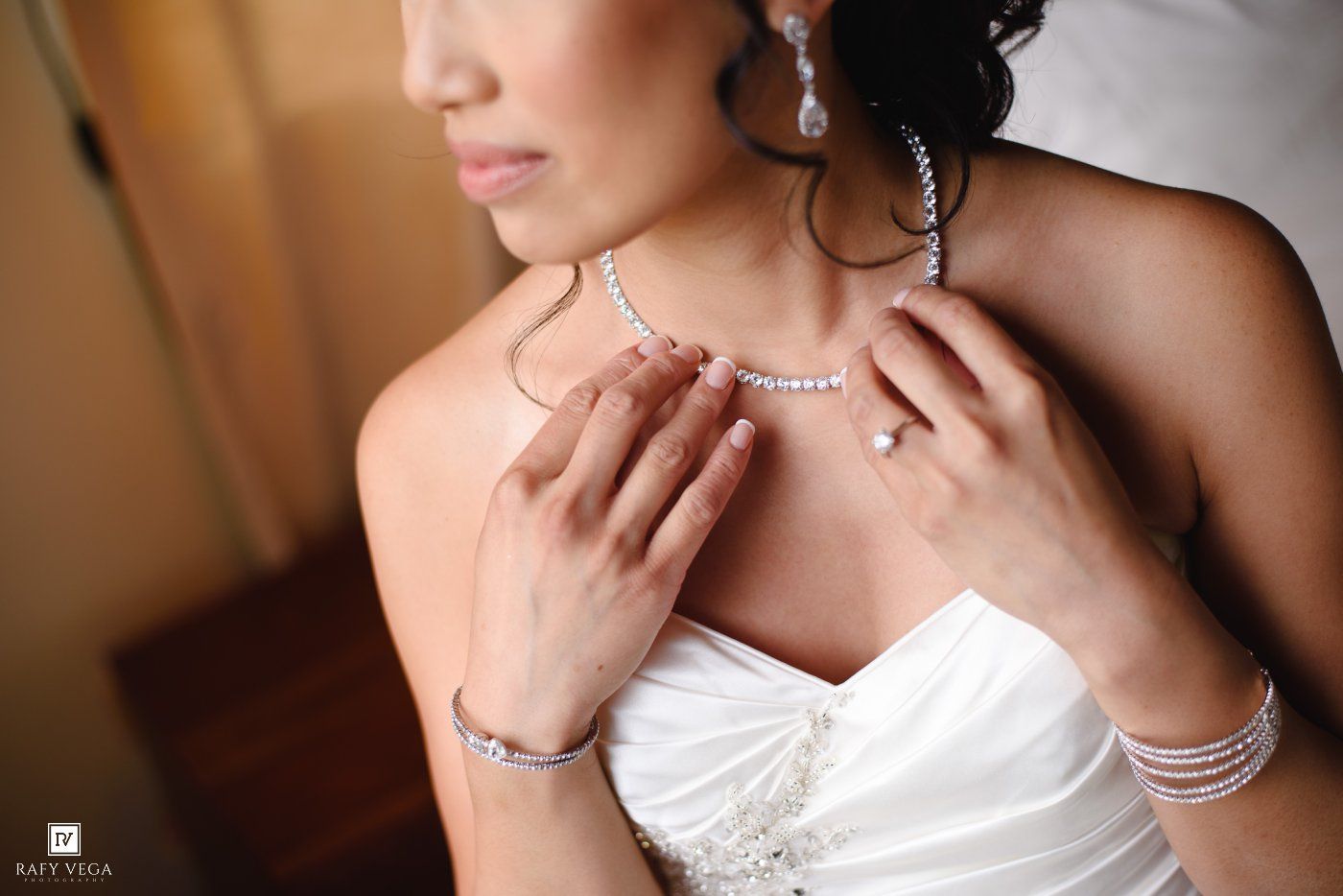 Bride adjusting diamond necklace wearing white wedding dress, bracelet and engagement ring on wedding day.
