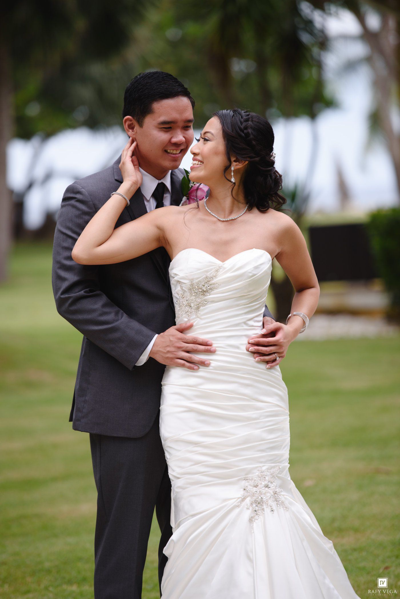 Bride and groom share a romantic embrace outdoors, she in a strapless mermaid gown and he in a gray suit.