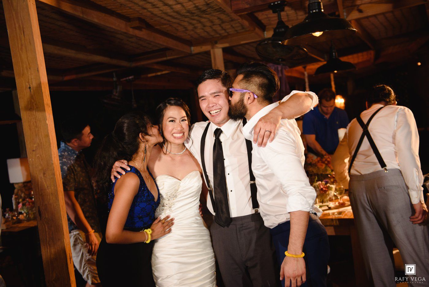 Bride and groom celebrate with a guest at their indoor wedding reception, sharing a joyful group hug.