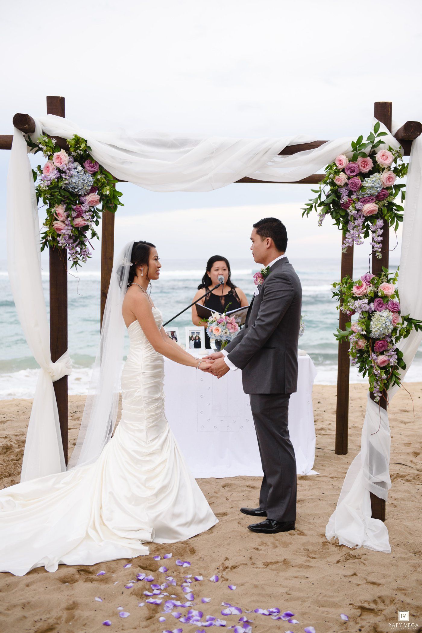 Bride and groom exchange vows under floral arch on beach wedding ceremony with officiant and ocean backdrop.