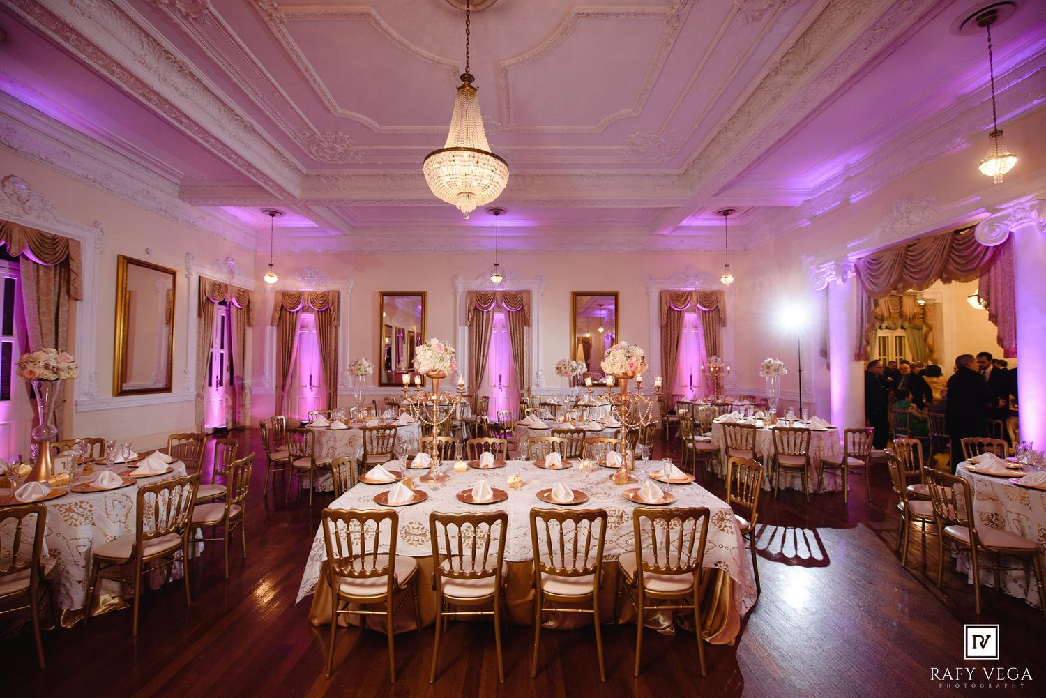 Elegant wedding reception hall with gold chairs, round tables, floral centerpieces, and romantic purple uplighting.