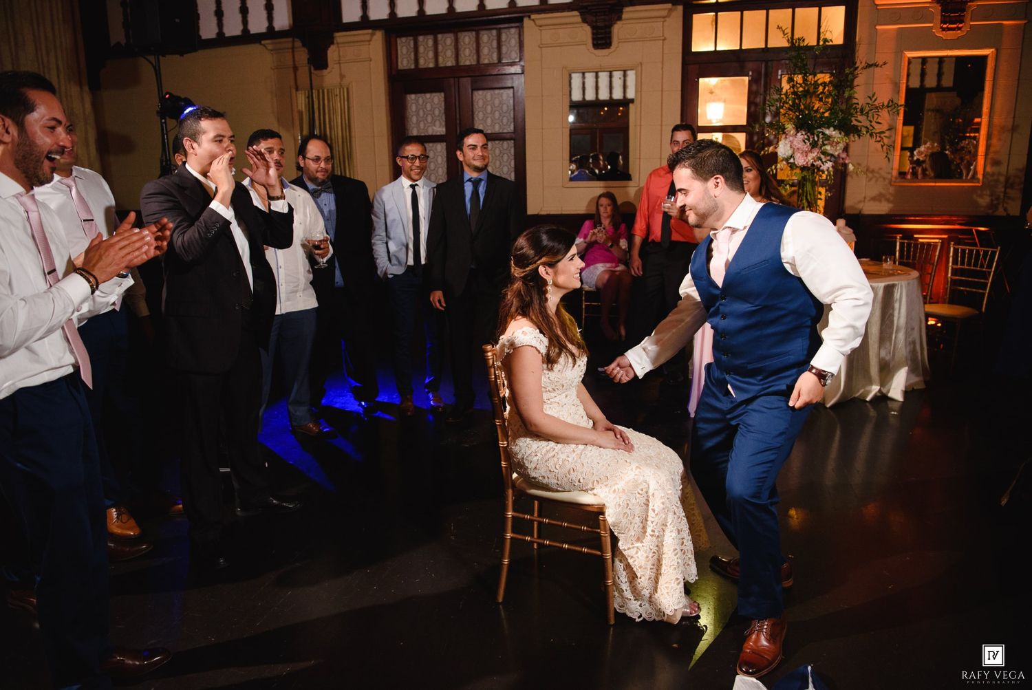 Bride seated on chair as groom dances at wedding reception while guests clap and celebrate in elegant venue.