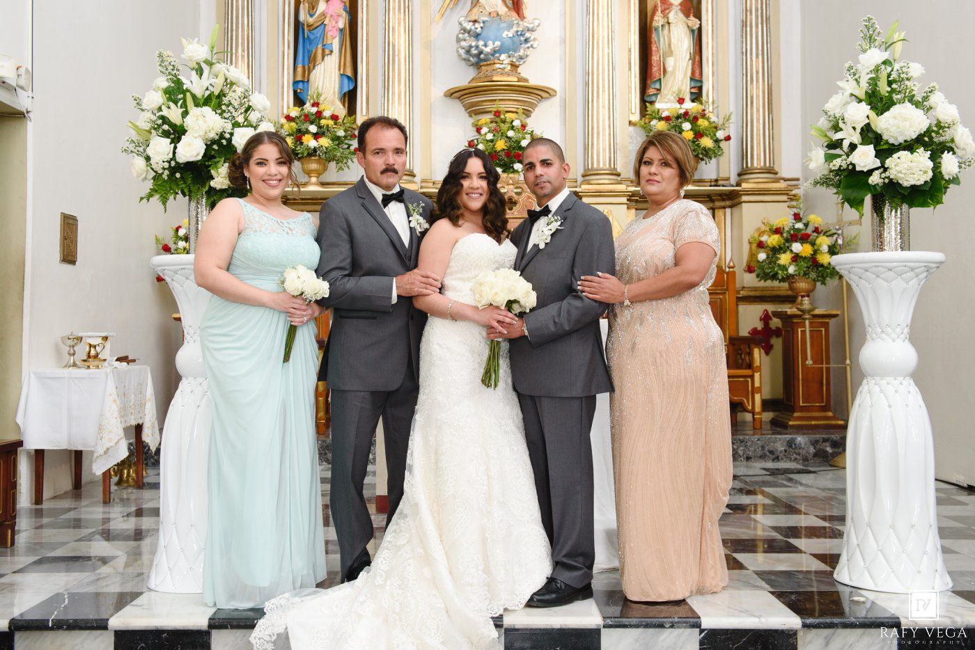 Wedding party posing in a church with white floral arrangements, bride in white gown flanked by family in formal attire.