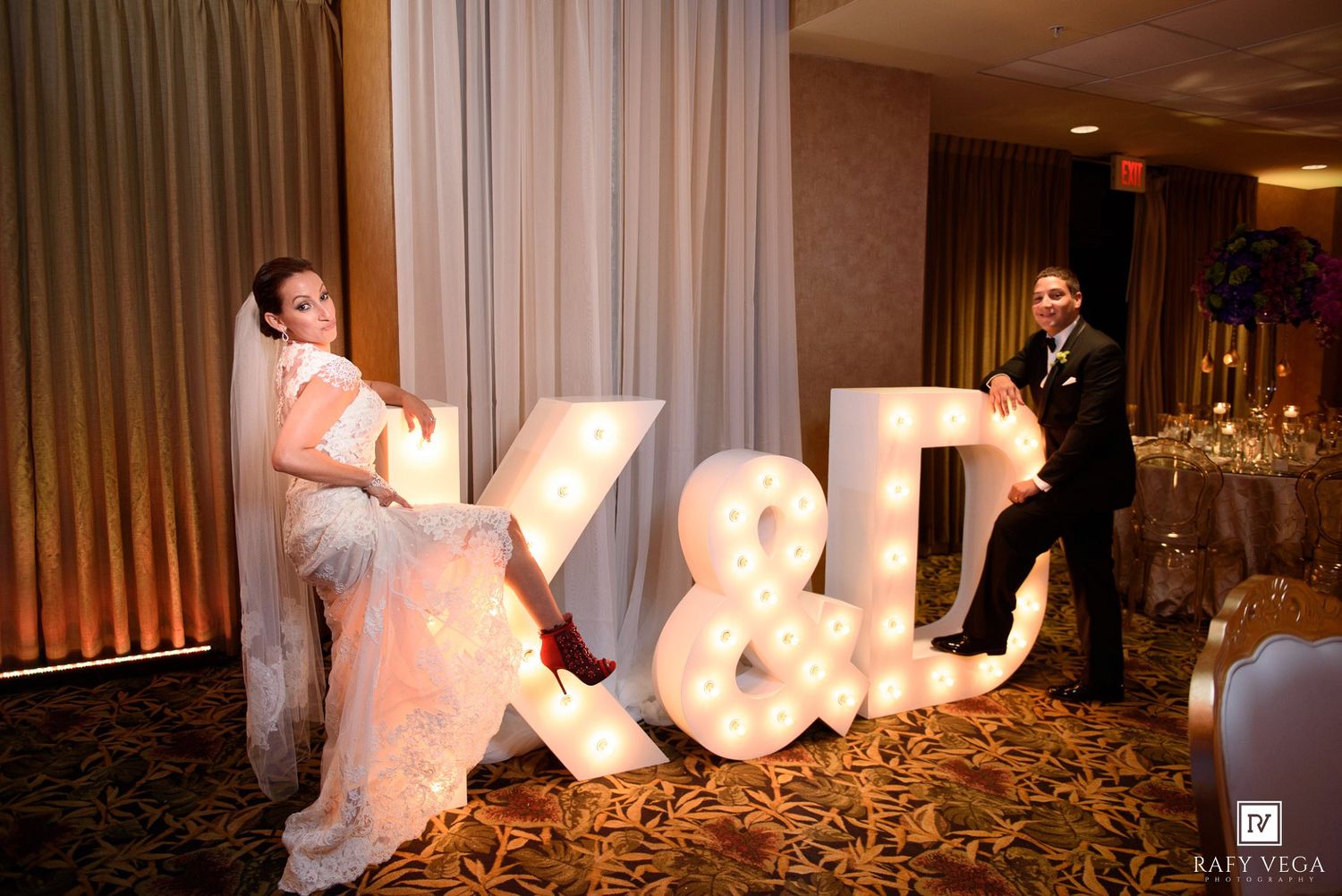 Bride and groom pose with illuminated marquee letters K&D at elegant wedding reception venue.