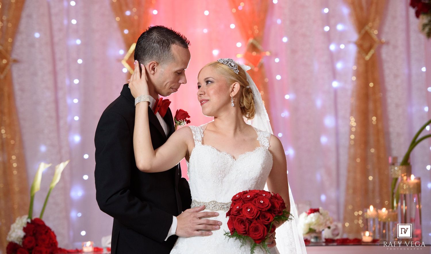 Bride and groom share a romantic moment at wedding reception with red roses and twinkling lights backdrop.