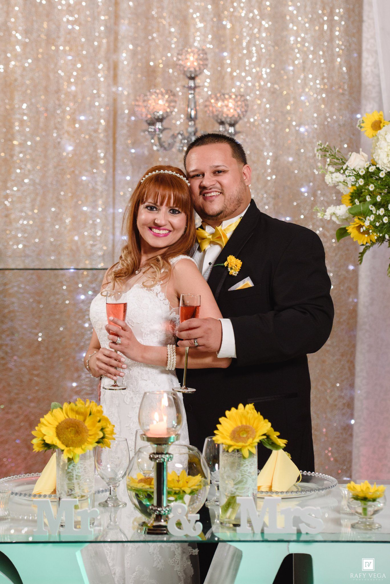 Bride and groom smiling at sweetheart table with sunflowers, holding champagne glasses at elegant wedding reception.
