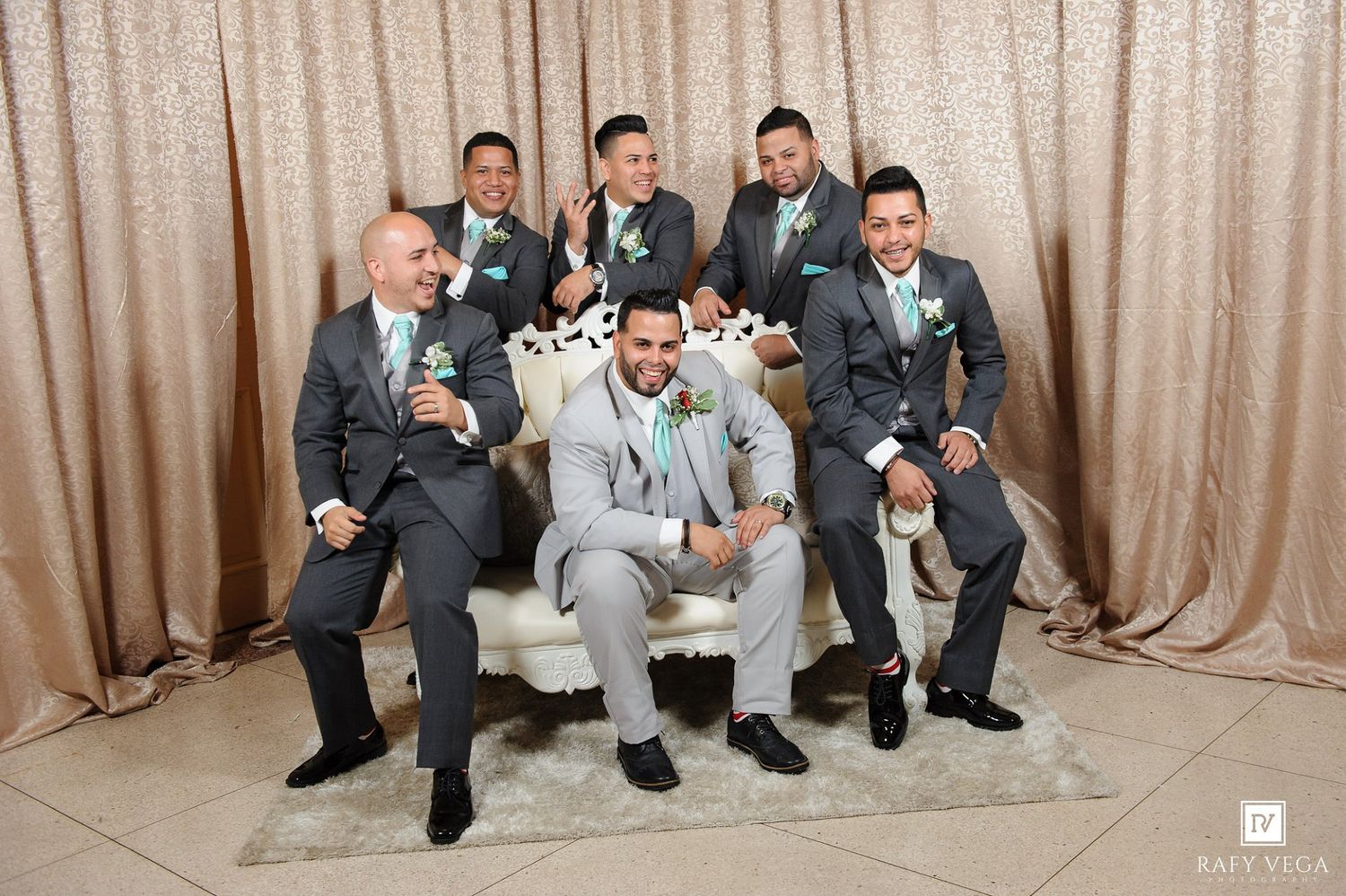 Groom in light gray suit sits on white chair surrounded by groomsmen in dark gray suits with mint boutonnieres.