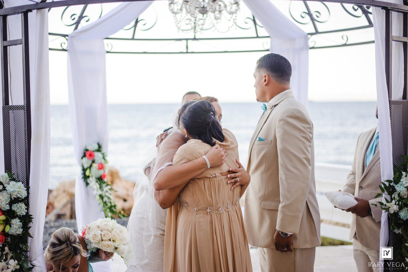 Bride sharing a heartfelt embrace with bridesmaid at beachside wedding ceremony under elegant gazebo.