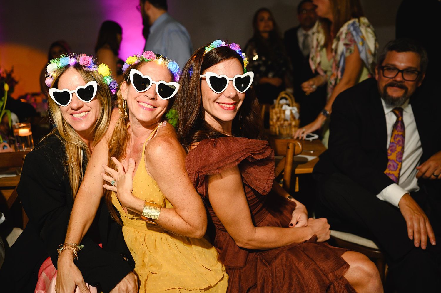 Three women wearing heart-shaped sunglasses and floral accessories pose together at a festive evening event.