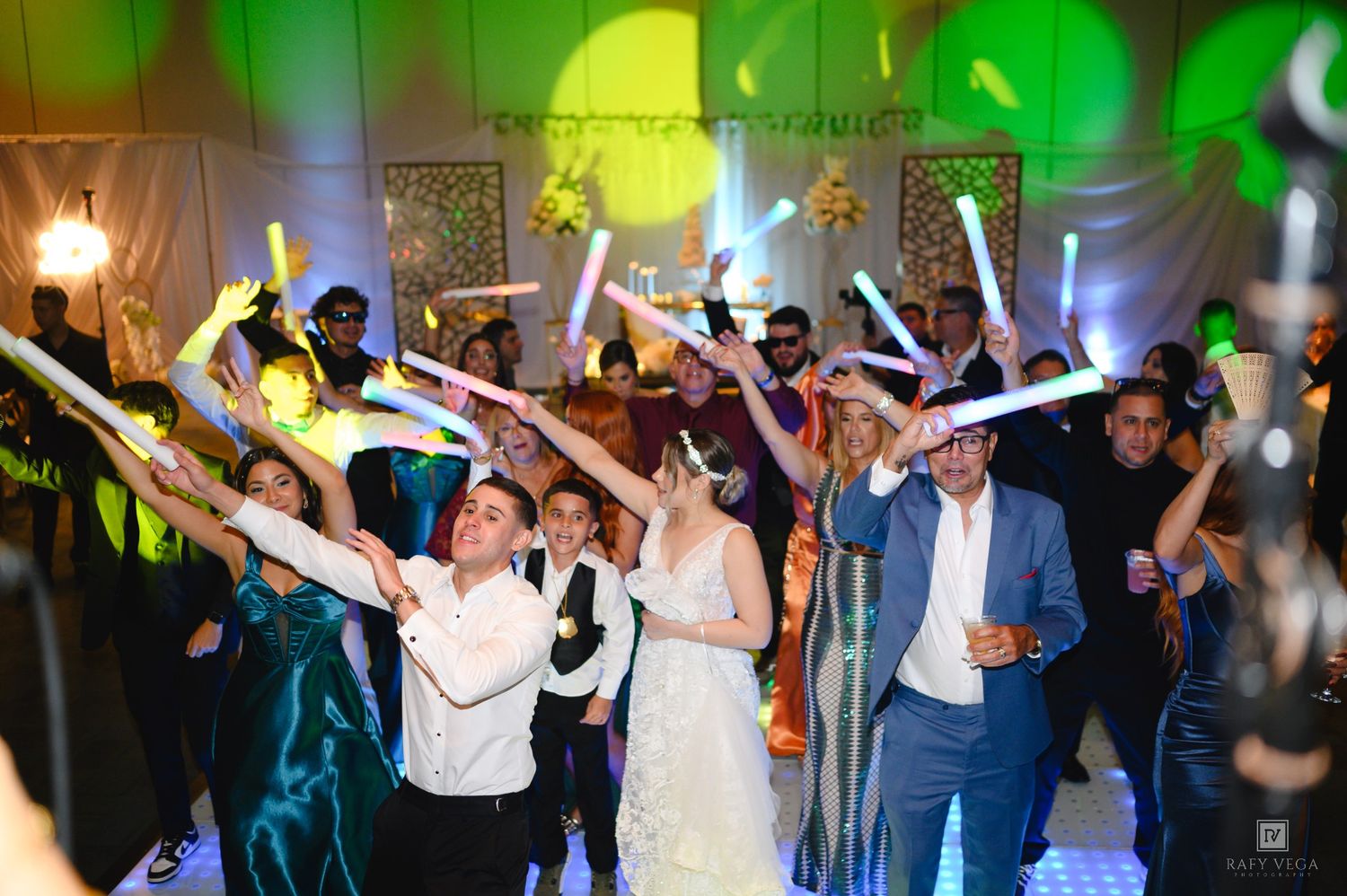 Wedding guests dance and wave colorful glow sticks during lively reception party celebration.