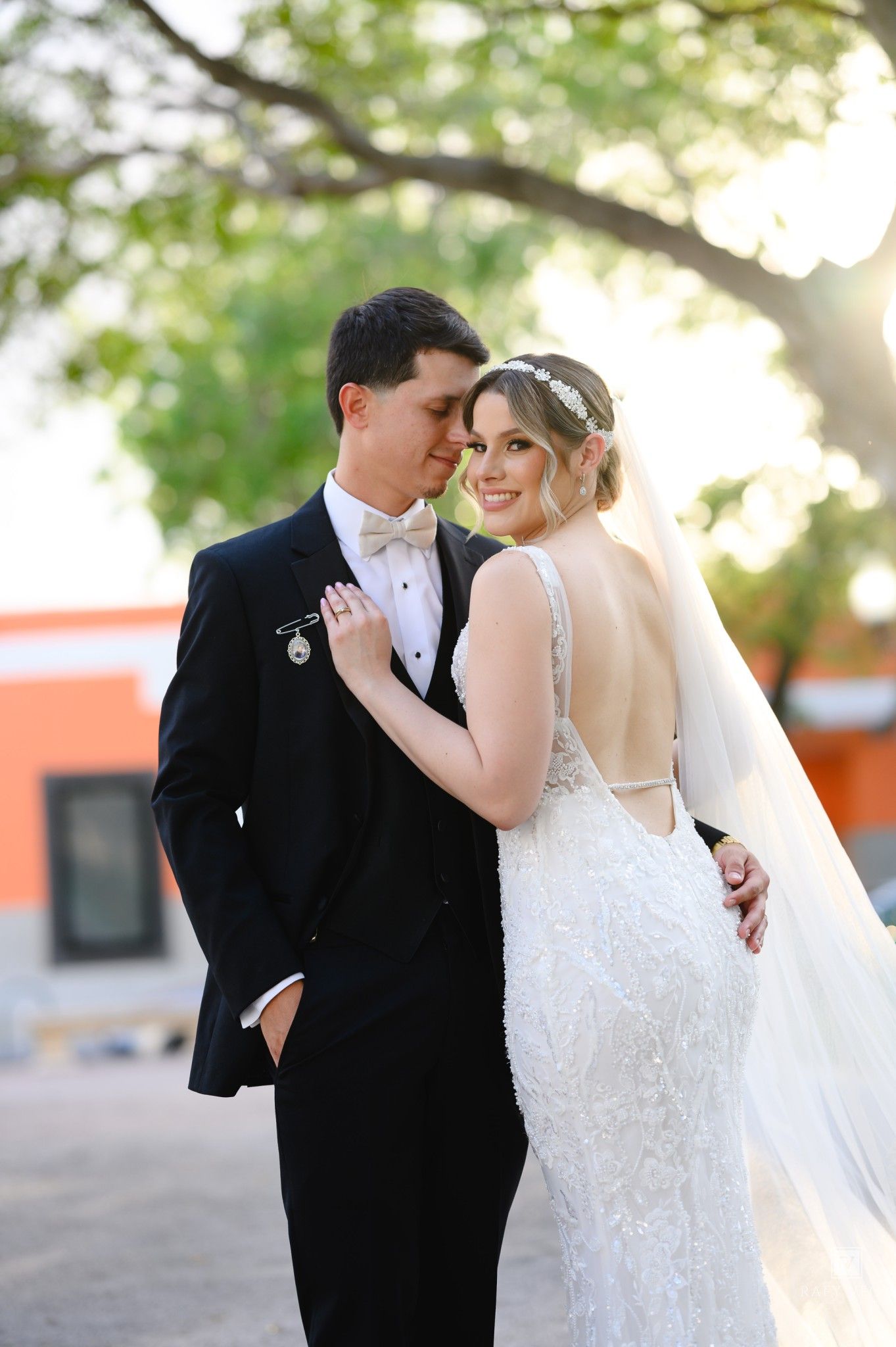 Elegant bride in backless lace gown and veil poses with groom in black tuxedo outdoors at wedding.