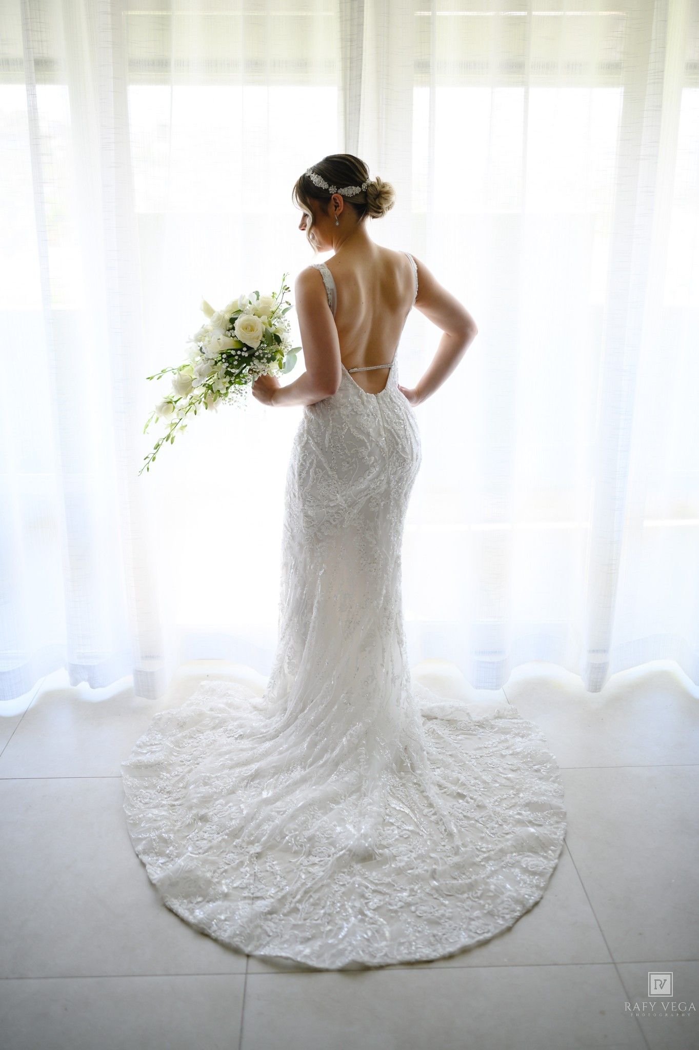 Bride in backless lace wedding gown holding white bouquet, standing by window with elegant train spread on floor.