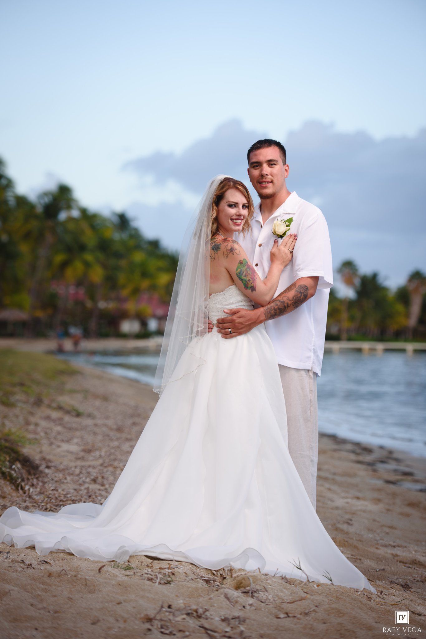 Bride and groom embrace on tropical beach, bride in white gown with veil, groom in white shirt, palm trees backdrop.