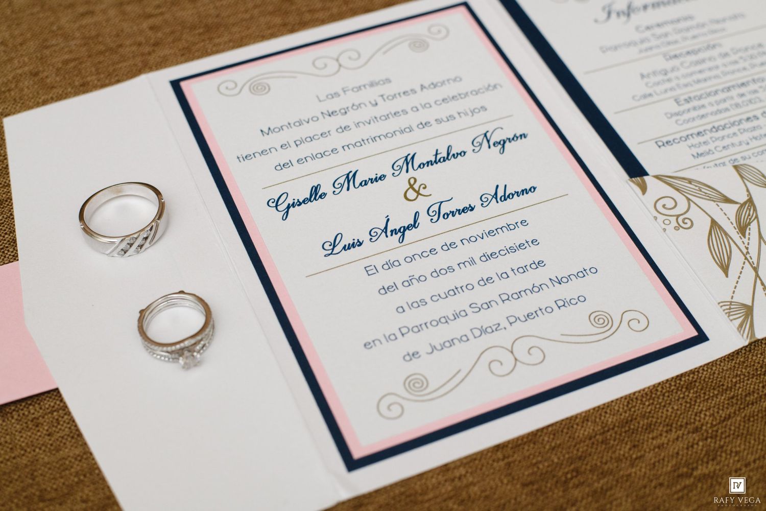 Elegant wedding invitation in Spanish with navy blue and pink accents, accompanied by two diamond engagement rings on a white surface.