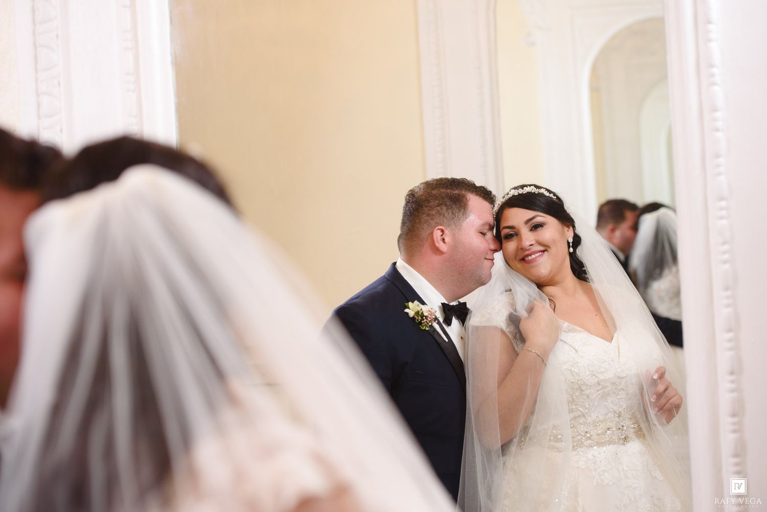 Groom kisses bride on cheek as she smiles at mirror reflection in beautiful white wedding gown and veil.