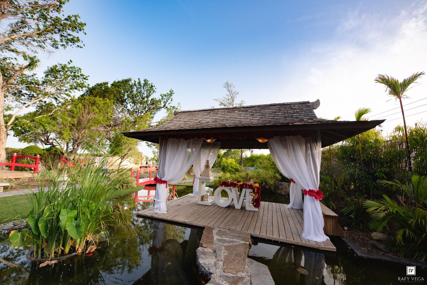Romantic outdoor gazebo with white drapes and LOVE letters on a wooden deck surrounded by a serene pond and lush tropical gardens.