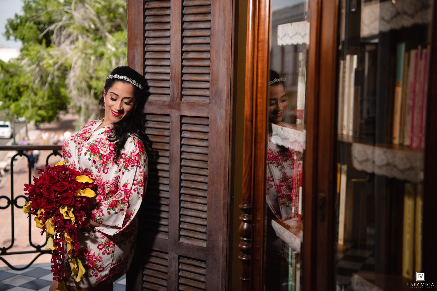 Bride in floral robe holding red bouquet stands by ornate wooden door, reflected in glass, on a sunlit balcony.