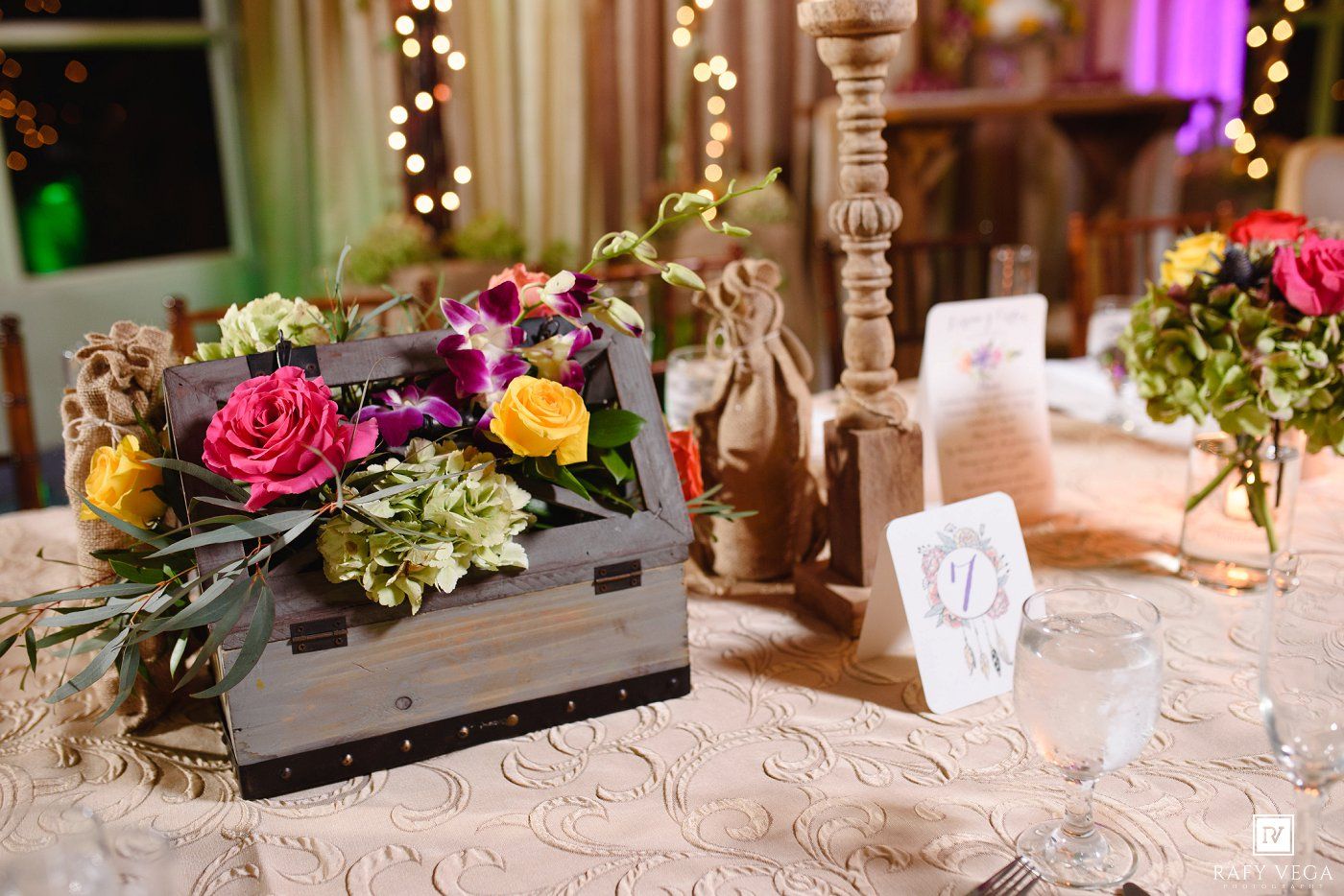 Rustic wooden crate centerpiece with colorful flowers including pink roses and yellow blooms on an elegant lace tablecloth.