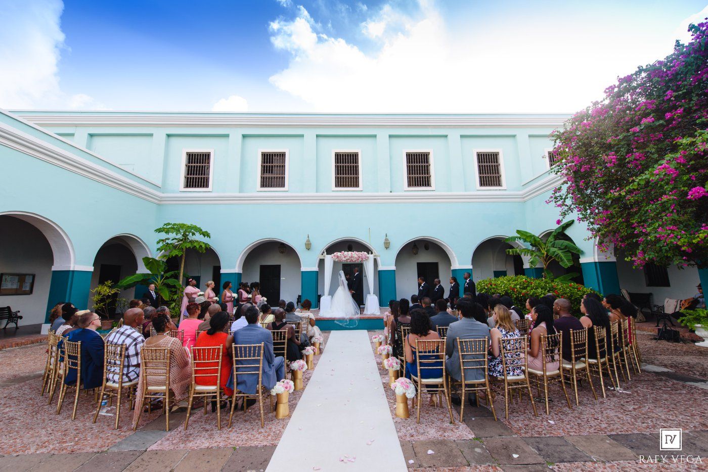 Outdoor wedding ceremony in a colorful courtyard with guests seated in chairs facing the altar under a blue sky.