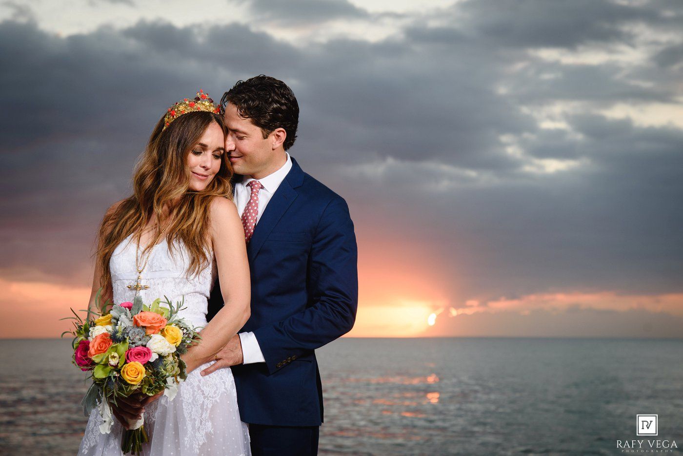 Bride and groom embrace at sunset over the ocean, she holds a colorful bouquet, wearing a floral crown, he in a navy suit.