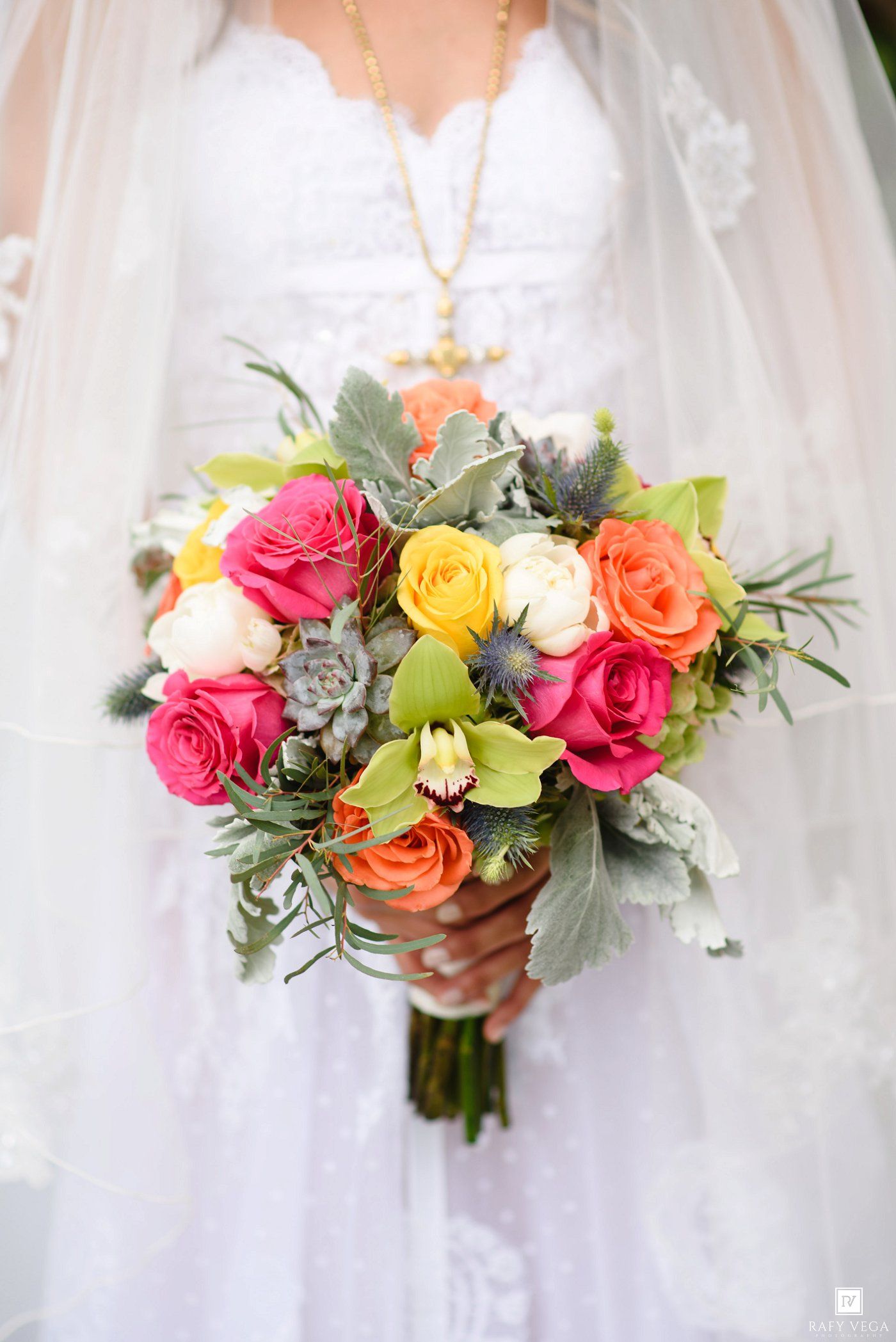 Bride in white lace gown holding vibrant wedding bouquet with pink, orange, yellow roses, green orchids, and silver foliage.