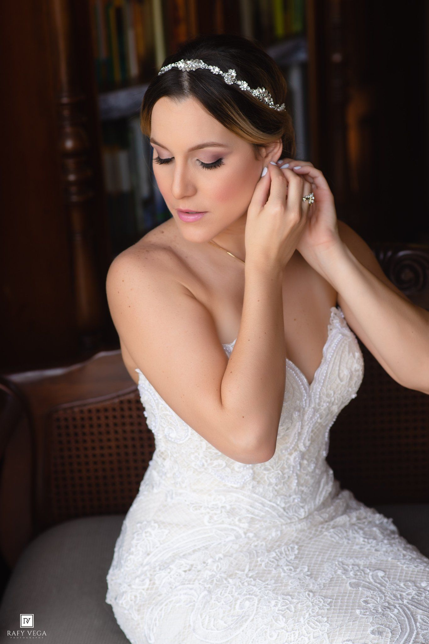 Bride in white lace gown and crystal headpiece putting on earrings while seated, wedding day getting ready photo.