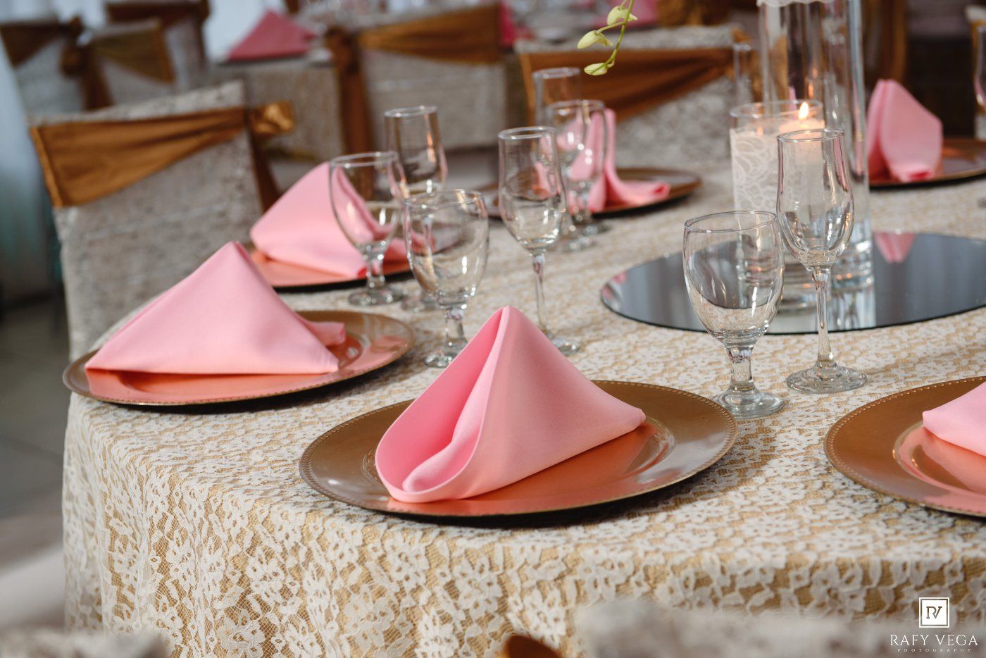 Elegant wedding reception table set with pink napkins, gold plates, crystal glasses, and candles on lace tablecloth.