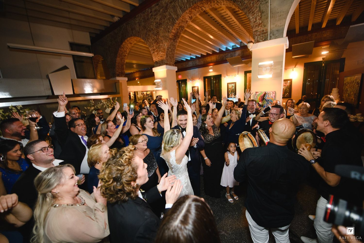 Joyful wedding reception crowd dancing and celebrating with bride in elegant venue with raised hands.