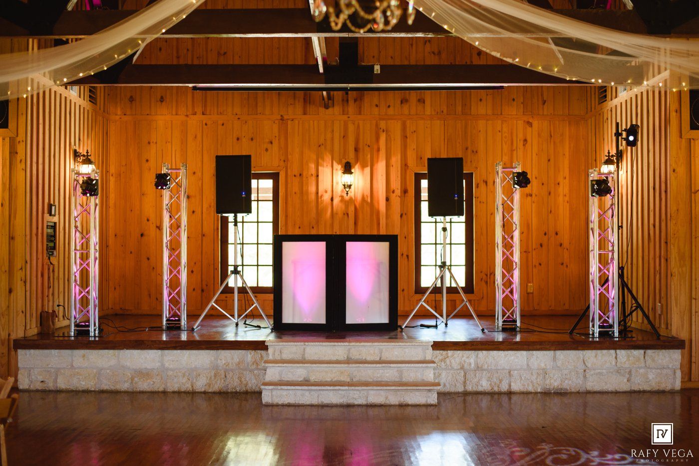 Rustic wooden barn wedding venue stage with pink illuminated DJ booth, speaker stands, and string lights for elegant receptions.