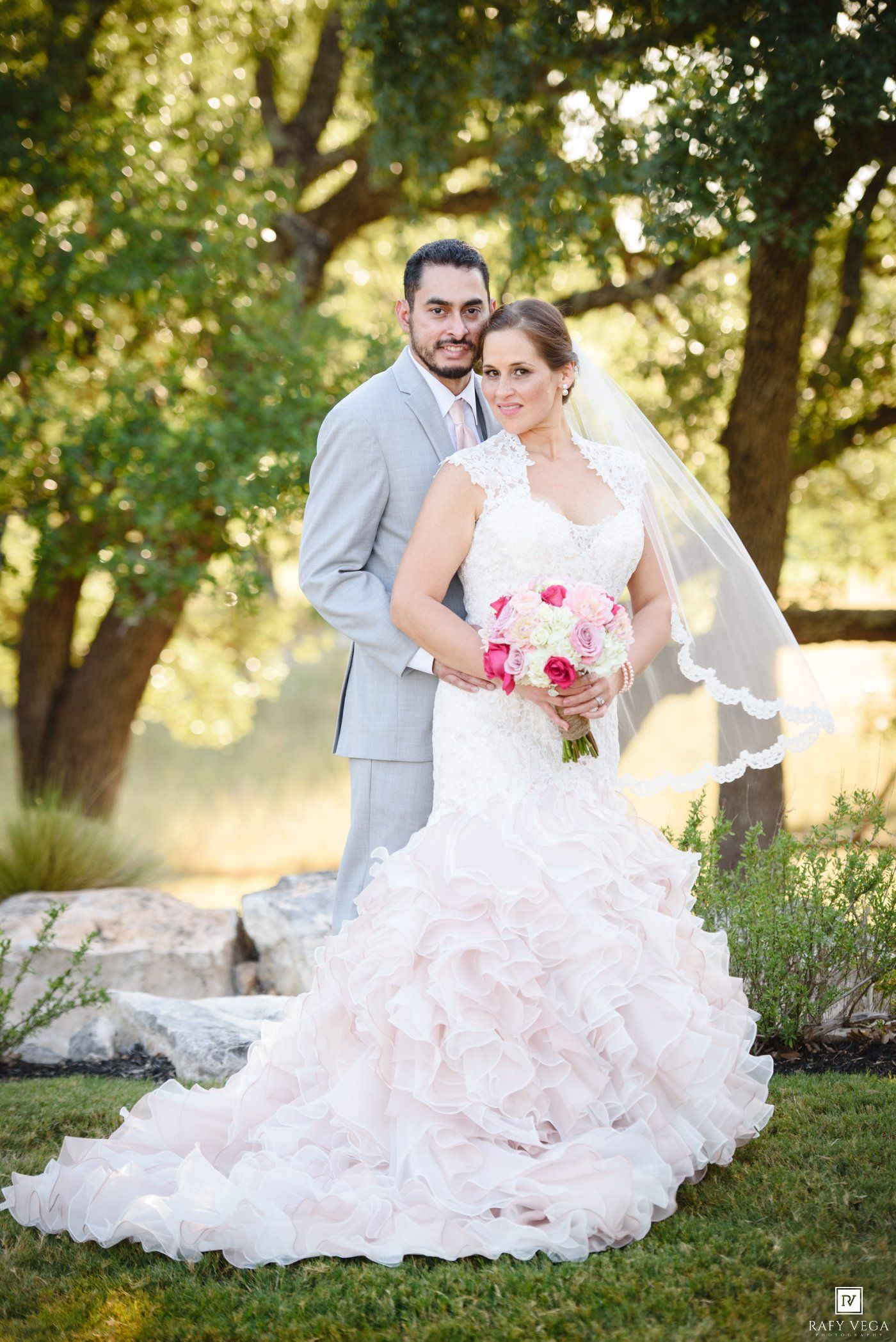 Bride in ruffled blush gown holding pink bouquet with groom in gray suit posing outdoors under green trees.
