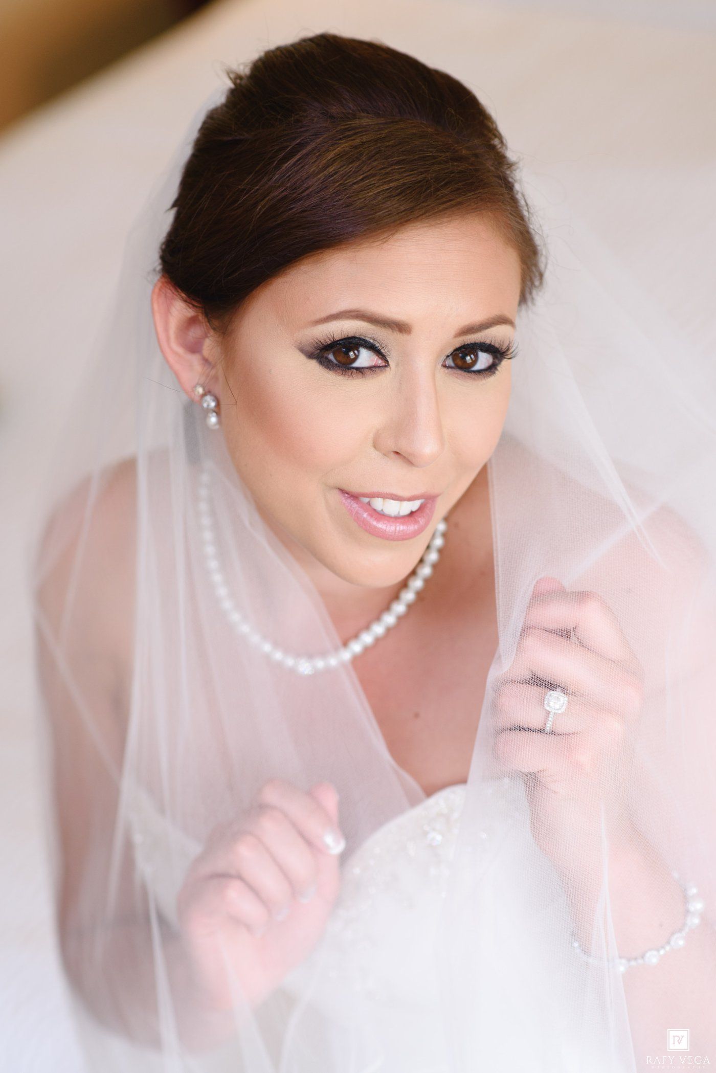 Bride in white wedding dress and veil wearing pearl necklace and diamond ring, with elegant bridal makeup.
