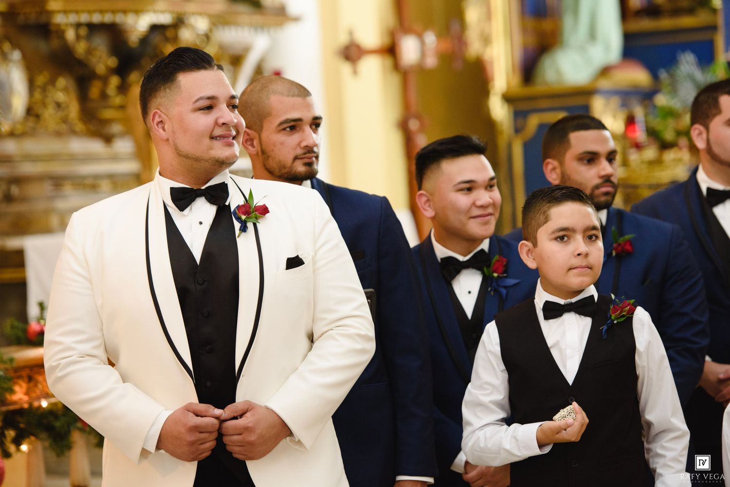 Groom in white tuxedo stands with groomsmen in black suits at church wedding ceremony.