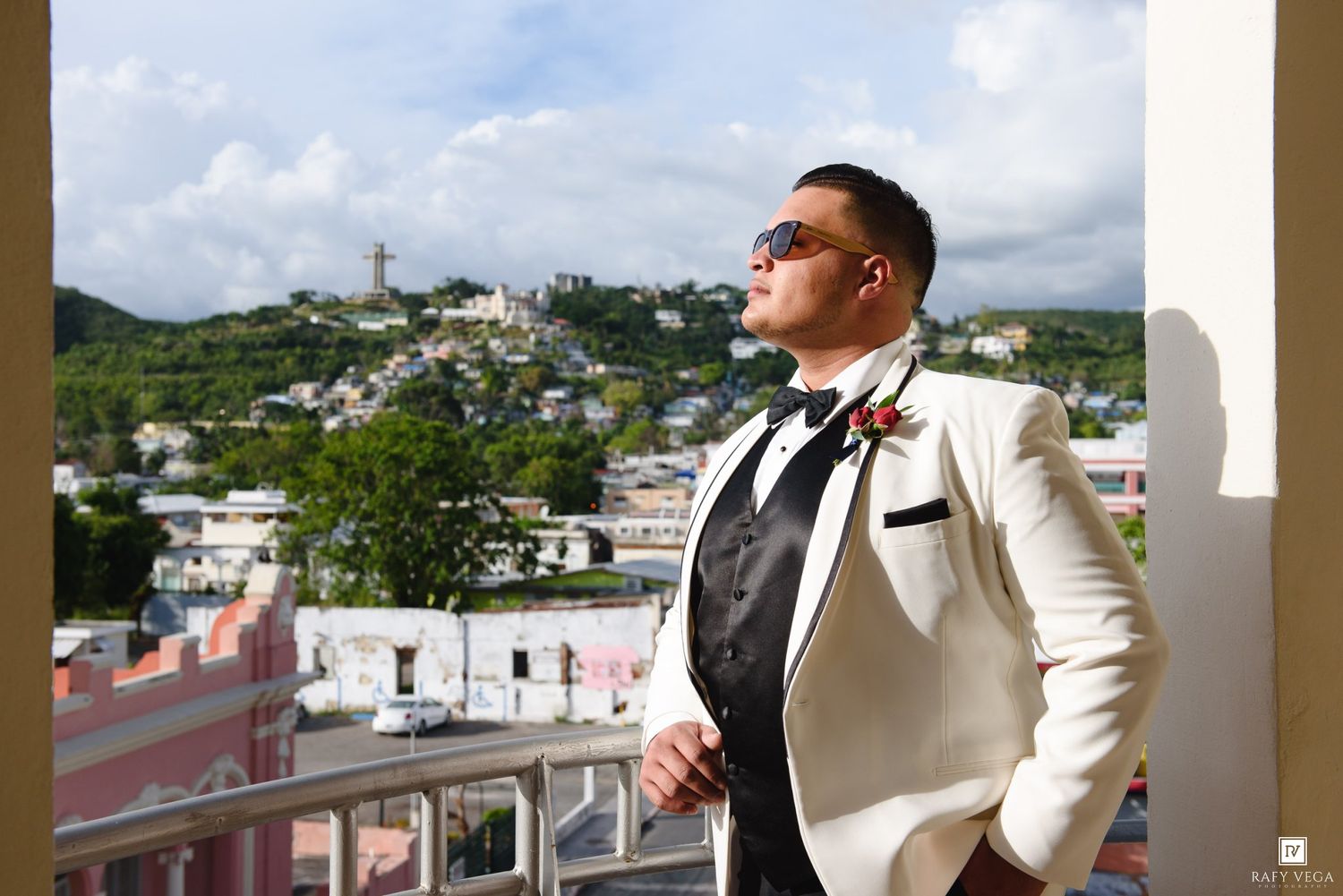 Groom in white tuxedo with bow tie poses on balcony overlooking colorful Caribbean town with lush green hills.