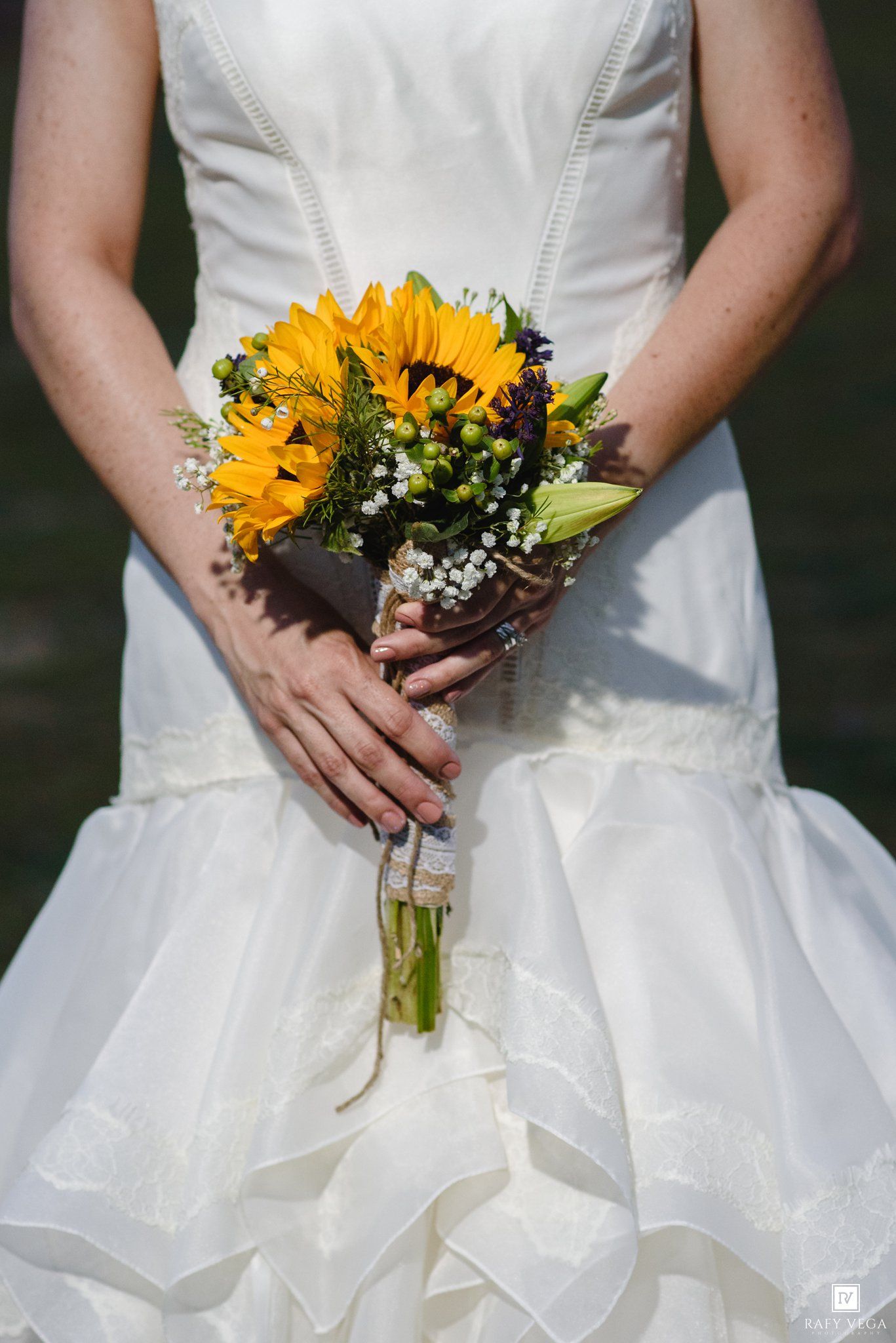 Bride in white wedding gown holding a vibrant sunflower bouquet with baby's breath at her wedding ceremony.