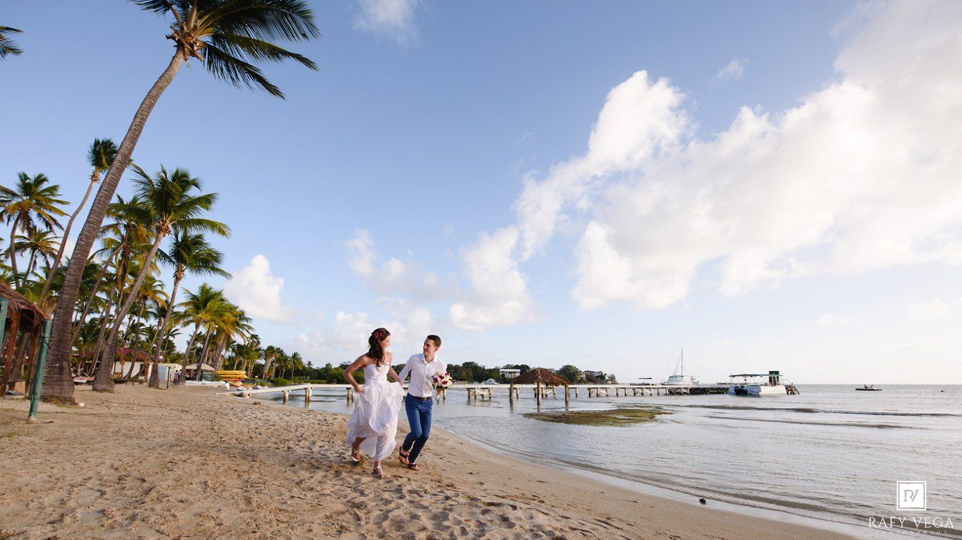 Couple walking hand in hand on a tropical beach with palm trees and a pier under a blue sky.