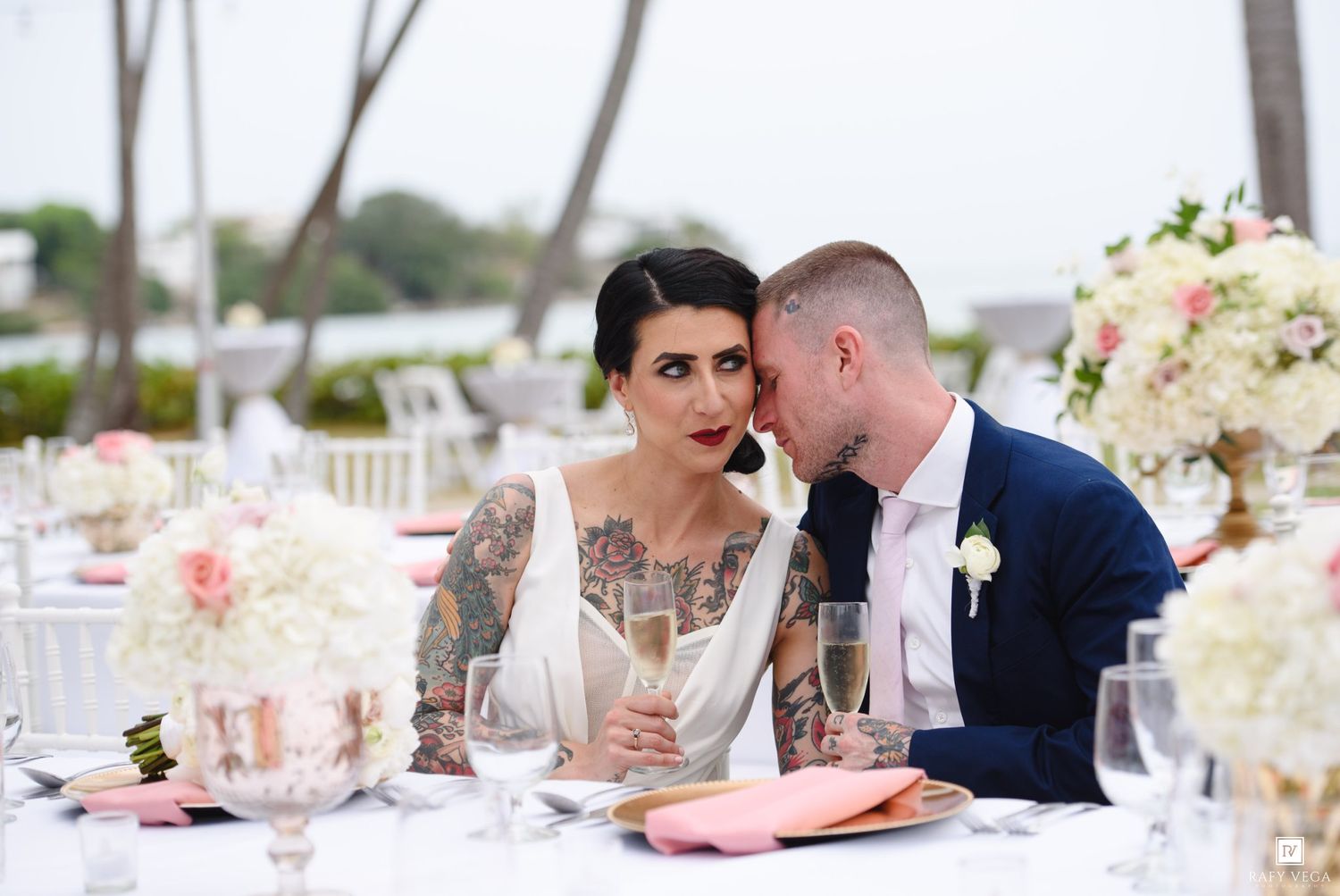 Tattooed bride and groom share intimate moment with champagne at wedding reception table.