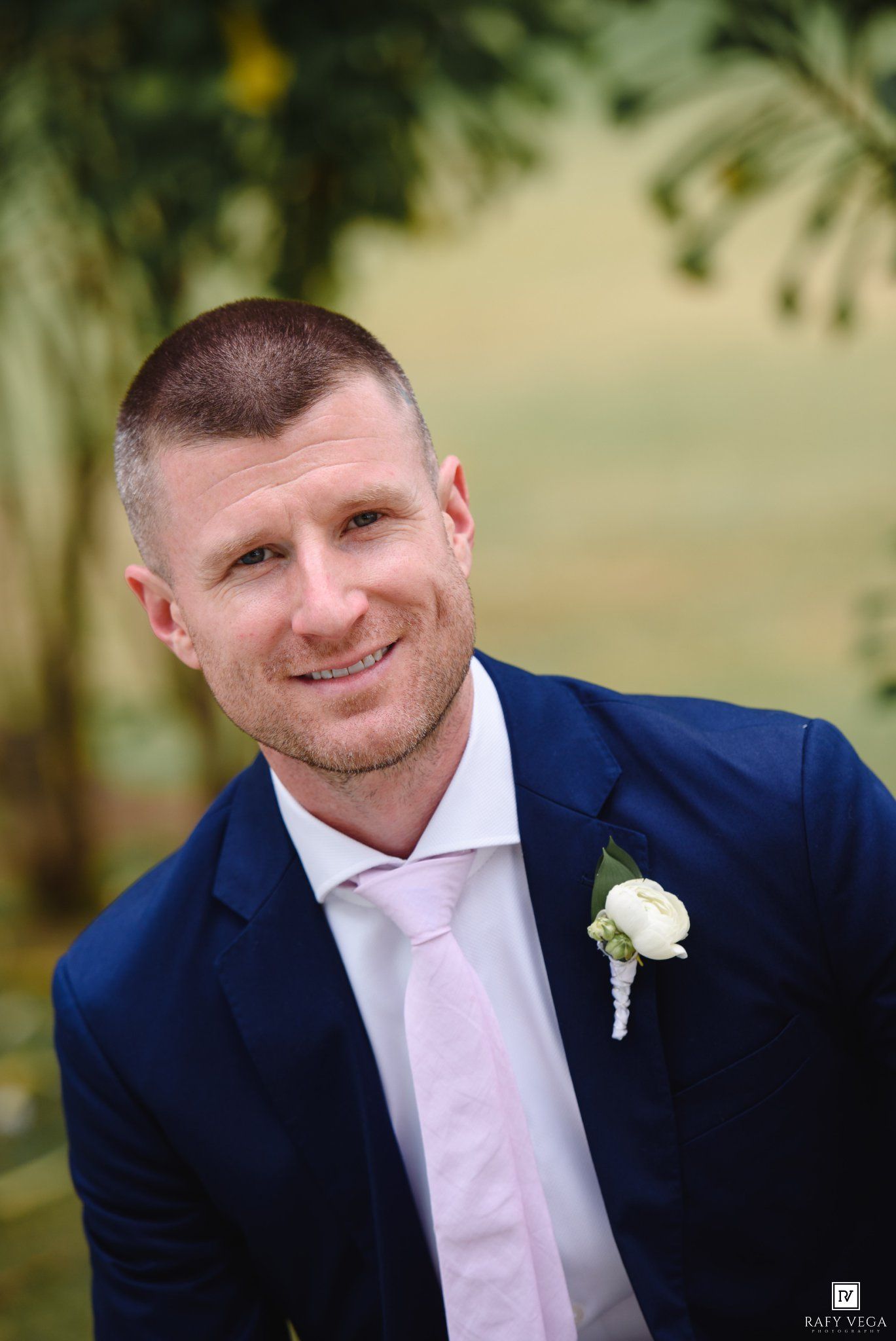 Smiling groom in navy blue suit with white boutonniere and pink tie poses outdoors at wedding.