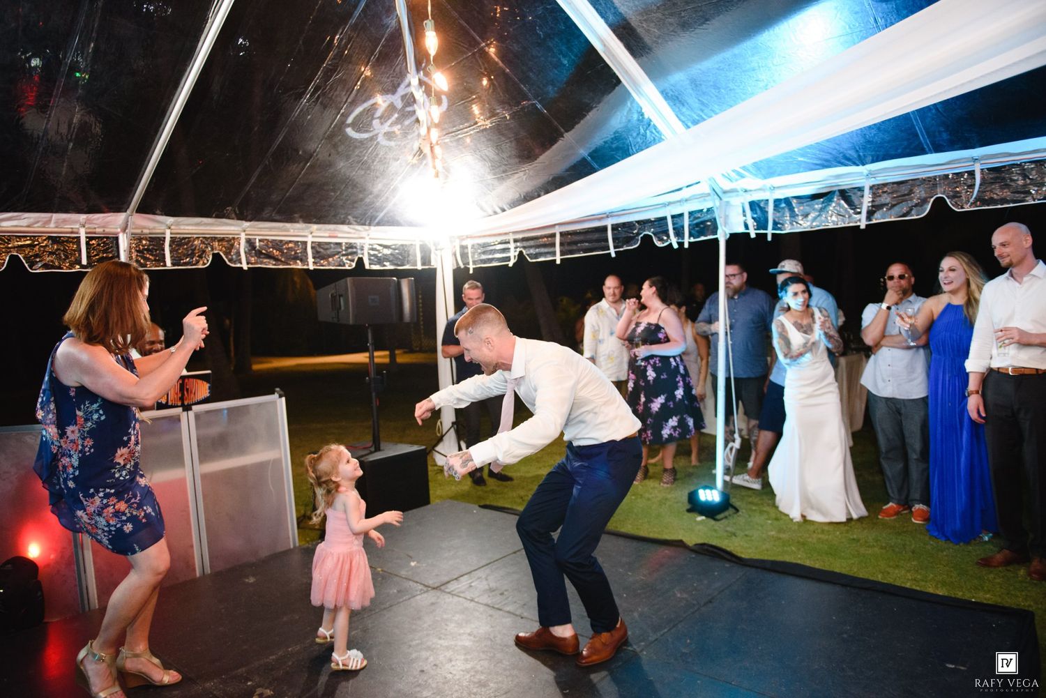 Groom dancing with a young girl in pink dress at outdoor wedding reception under a tent.