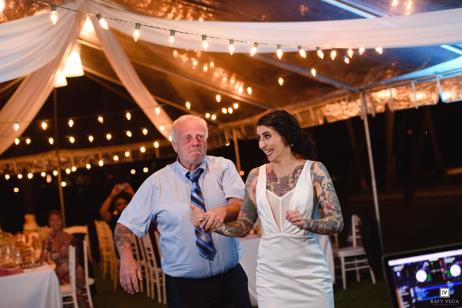 Bride with tattoos in white wedding dress walks with elderly man under string lights at outdoor reception.