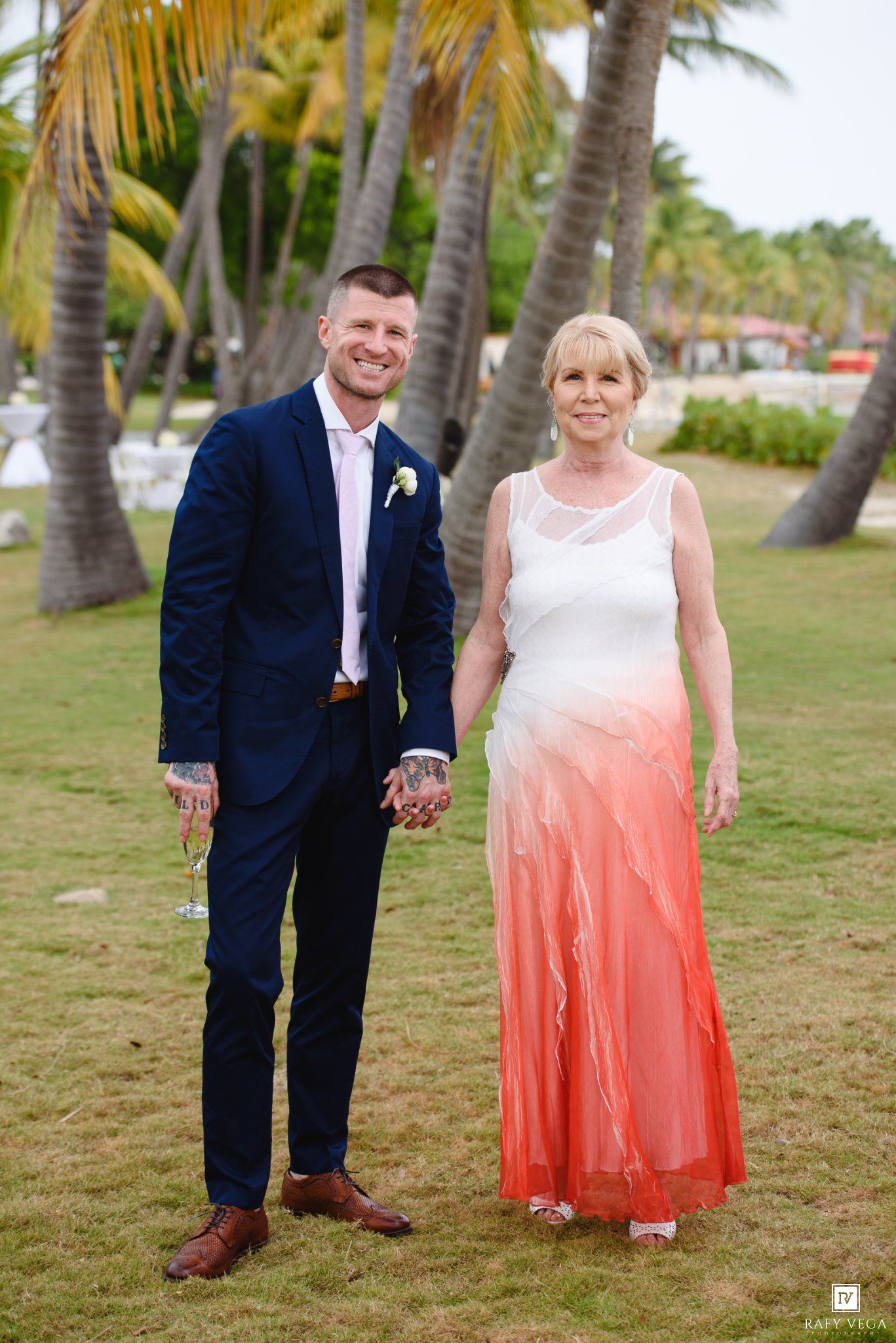 Groom in navy suit and woman in coral ombre gown pose together at tropical beach wedding venue.
