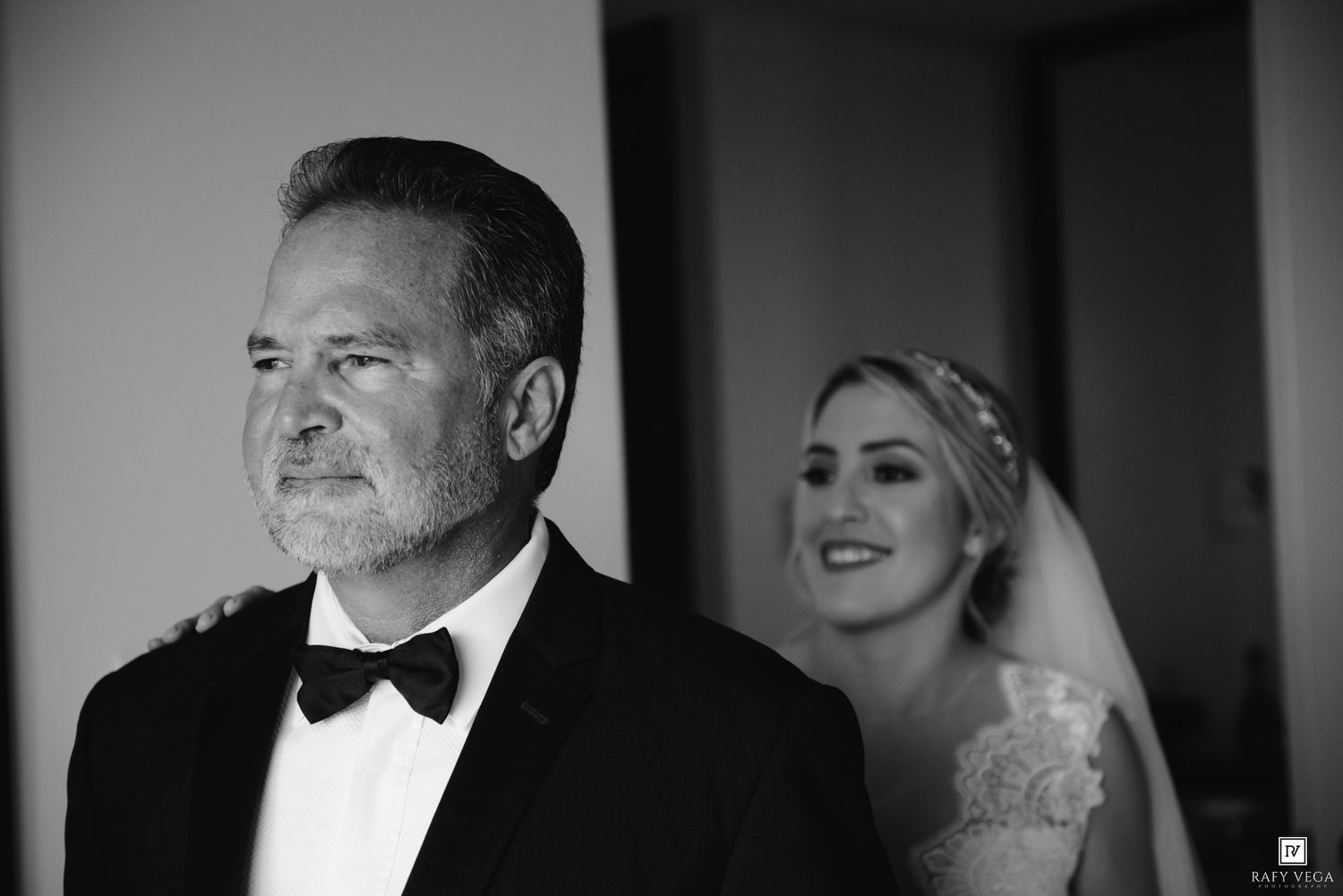 Black and white photo of a bride in veil smiling behind a man in tuxedo during an emotional wedding first look.