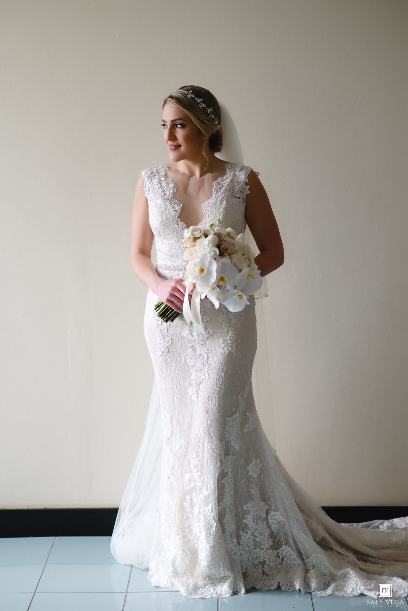 Bride in lace mermaid wedding gown holding white orchid bouquet, wearing crystal headpiece, posing elegantly indoors.