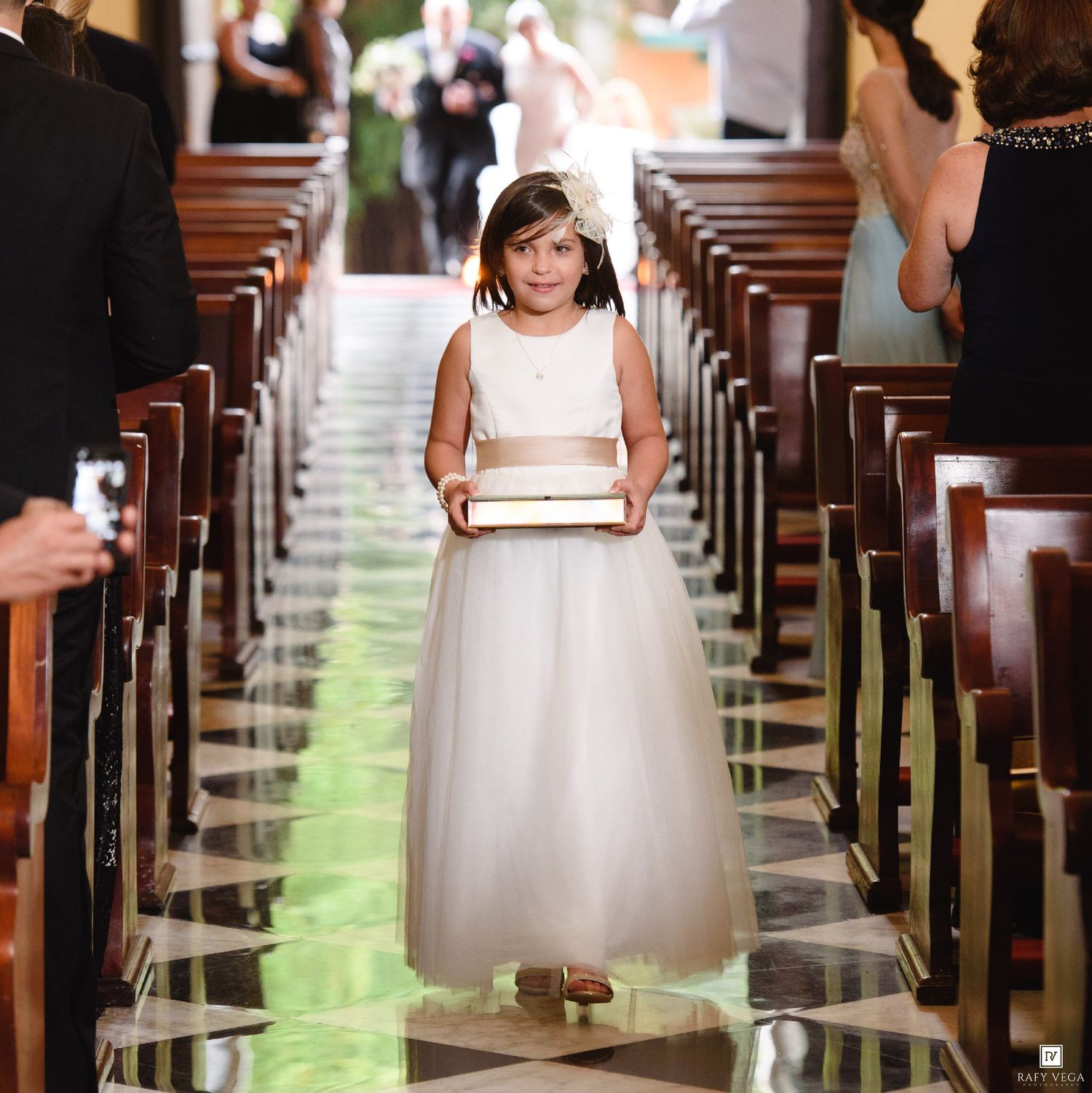 A young flower girl in a white dress walks down the church aisle carrying a decorative box at a wedding ceremony.