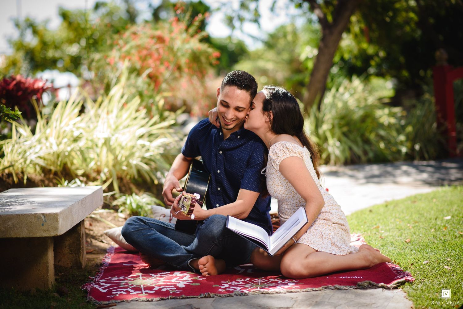 Couple sitting on a red blanket outdoors, woman kissing man's cheek as he plays guitar in a lush garden.
